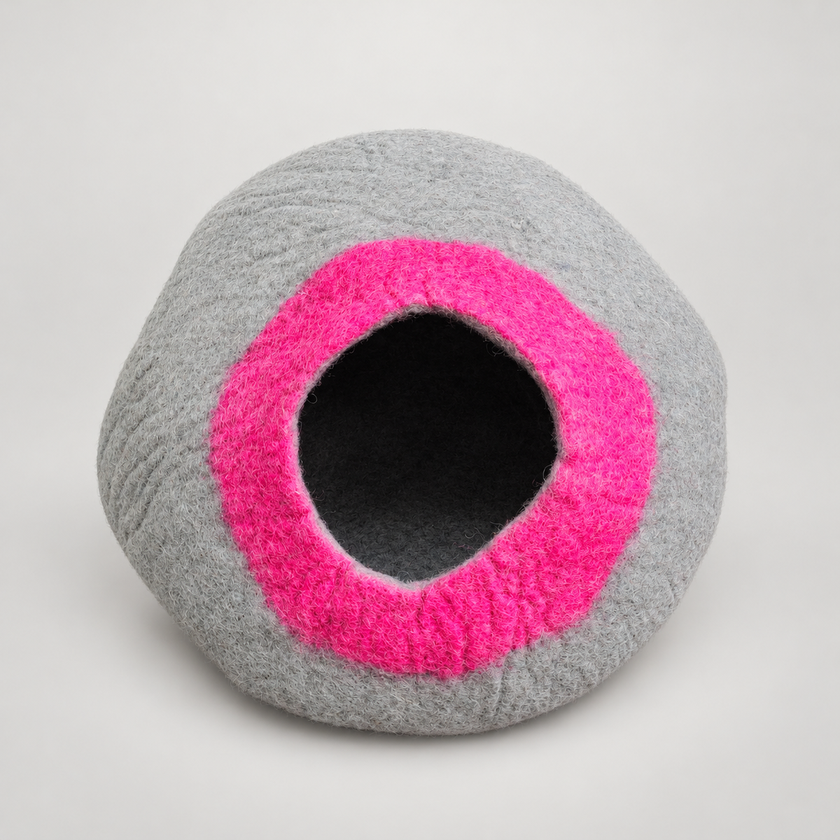Gray handmade wool cat cave with bright pink entrance on warm gray background.