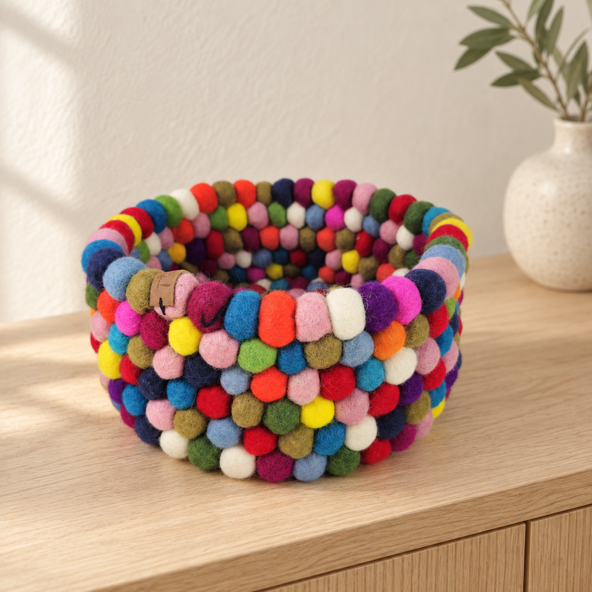 Multicolor felt ball basket styled on a pale wood console in a neutral interior.
