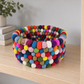 Round multicolor felt ball basket displayed on a light wood shelf beside neutral books.
