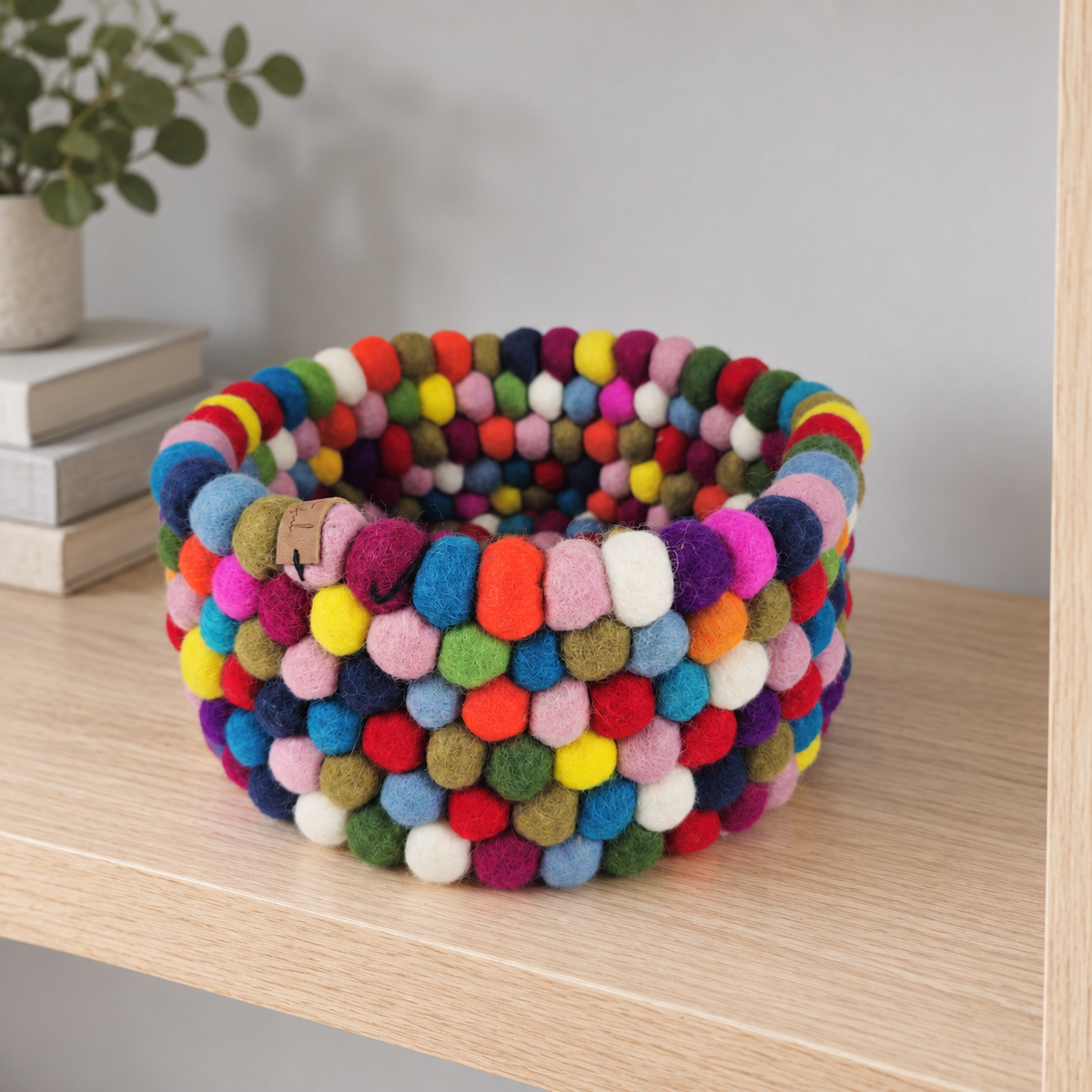 Round multicolor felt ball basket displayed on a light wood shelf beside neutral books.